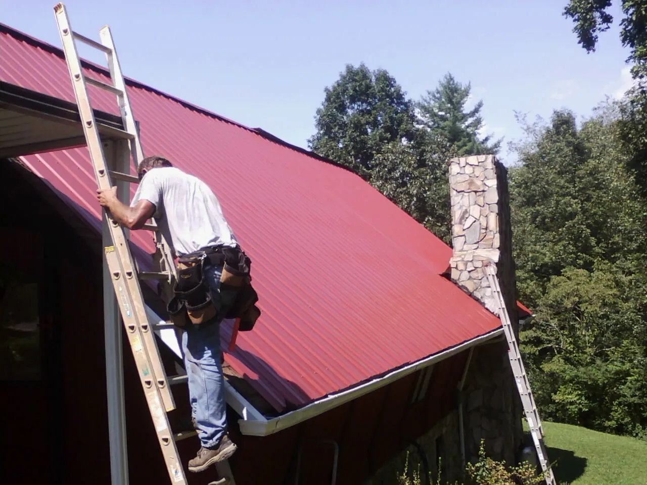 Roofer on a ladder beside a red metal roof and stone chimney — Soffit & Fascia Repair by Roofing Brothers in Courtenay
